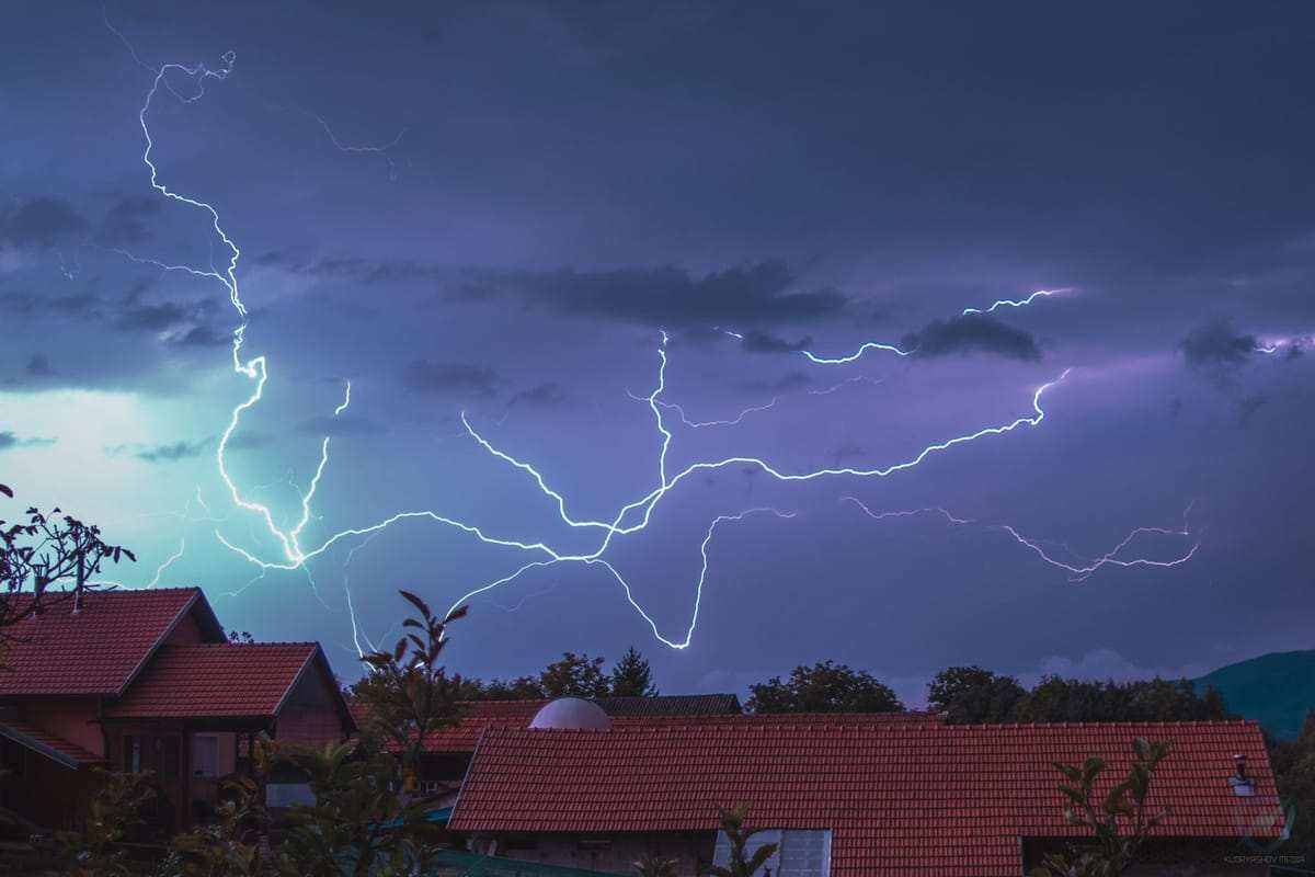 Žaibai virš namų raudonų čerpių stogais per perkūniją